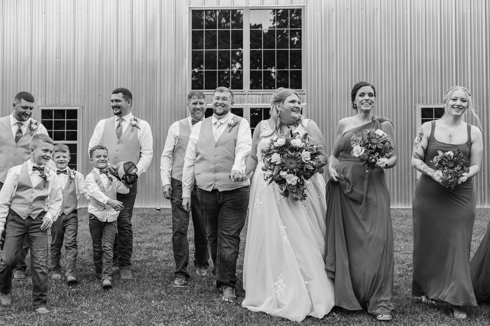 Wedding party walks together outside barn venue in black and white photo with bride, groom, and attendants.
