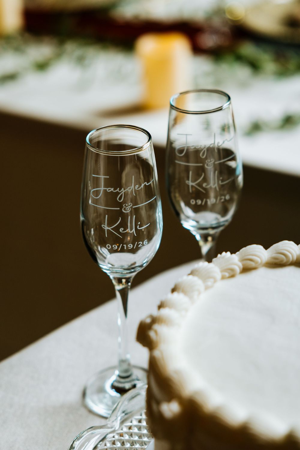 Custom engraved champagne flutes reading Jayden and Kelli 09/19/26 beside wedding cake on table.