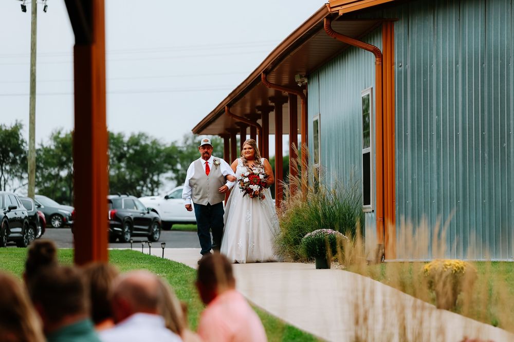 Bride walks down outdoor aisle with escort beside rustic barn wedding venue on sunny day.