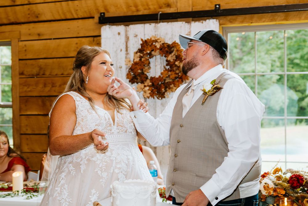 Bride playfully smears cake on grooms face during wedding reception in rustic wood barn venue.