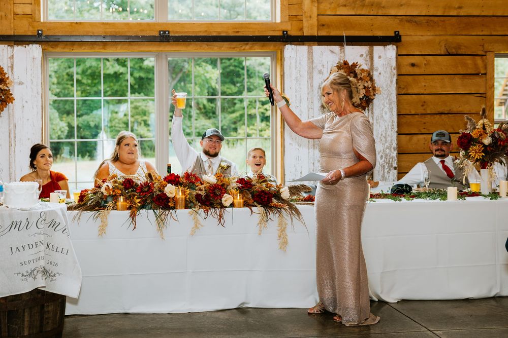 Woman gives toast at wedding reception as bride and groom sit at decorated head table.
