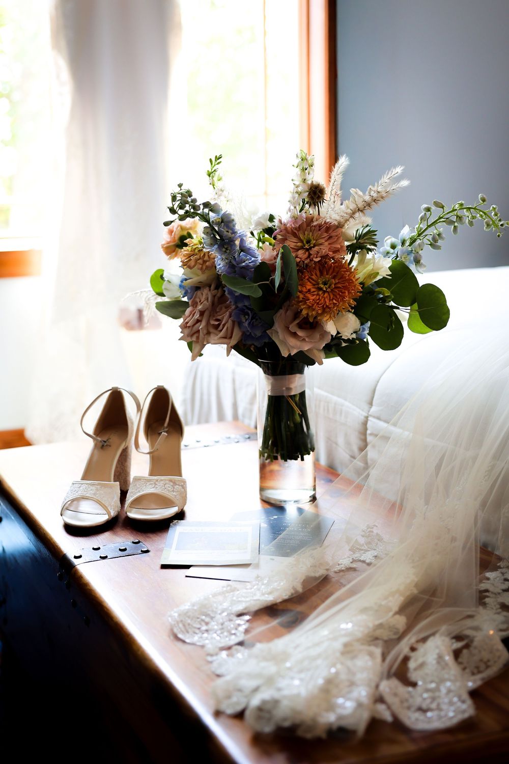 Bridal details flat lay with bouquet, heels, veil, and accessories arranged on wooden dresser by window.