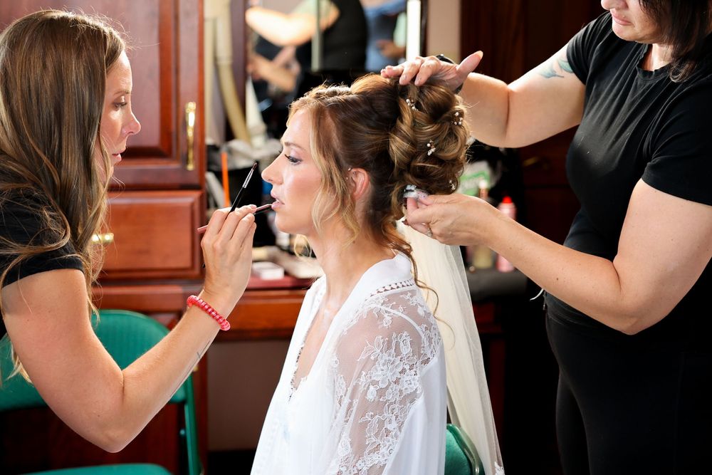 Two stylists apply makeup and style hair of bride in white robe before wedding ceremony.