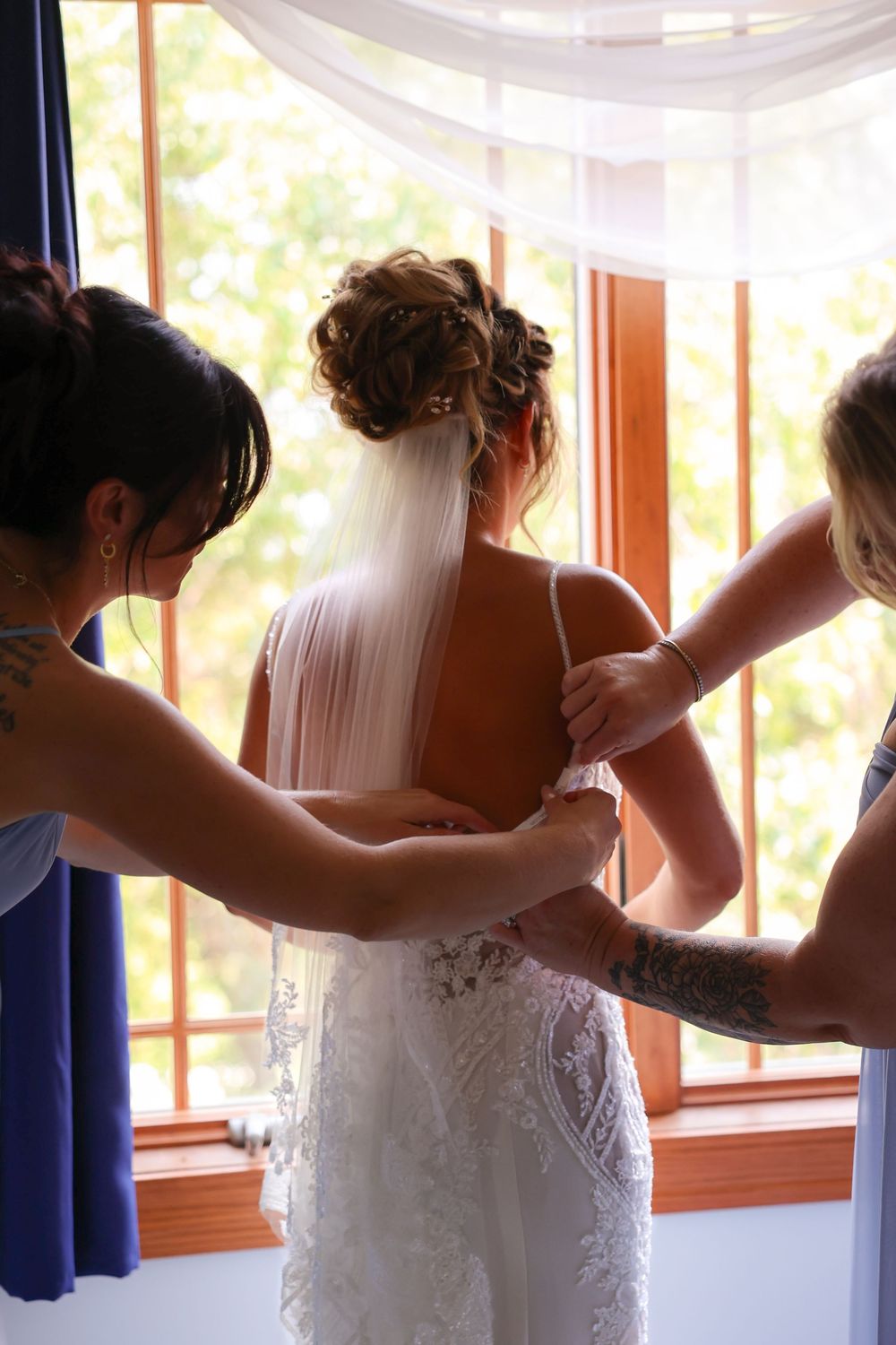 Two bridesmaids help fasten brides lace wedding gown near sunlit window before ceremony.