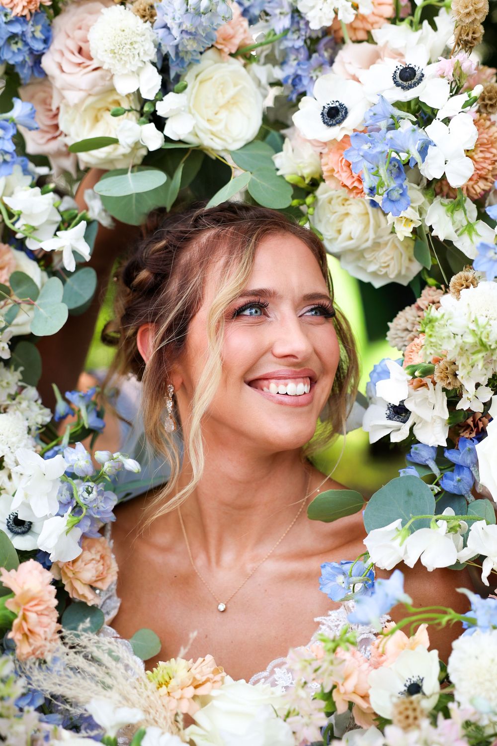 Smiling bride surrounded by blue, white, and peach floral arrangement looking up joyfully on wedding day.