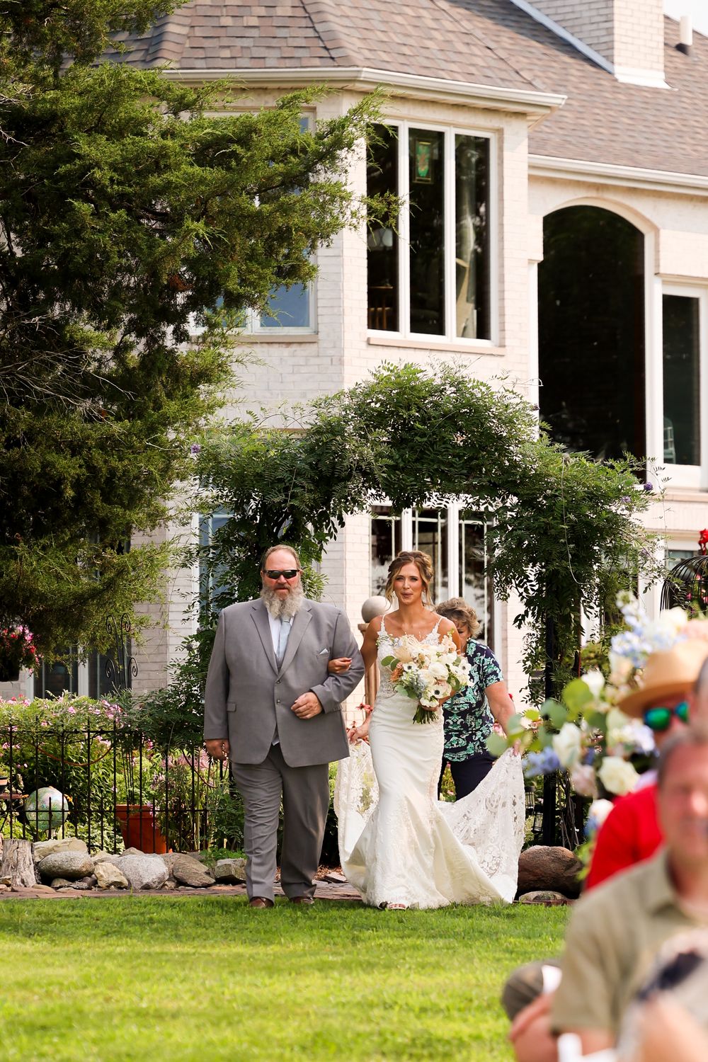 Bride in white gown walks with two escorts across green lawn toward outdoor wedding ceremony.