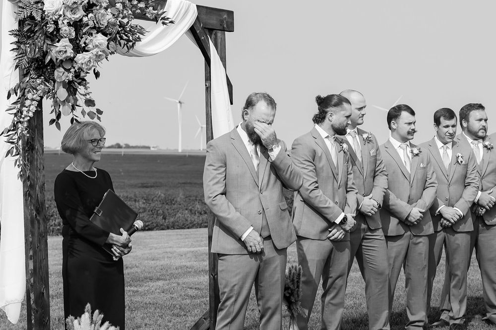 Groom emotional at altar with groomsmen in gray suits at outdoor farm wedding ceremony.
