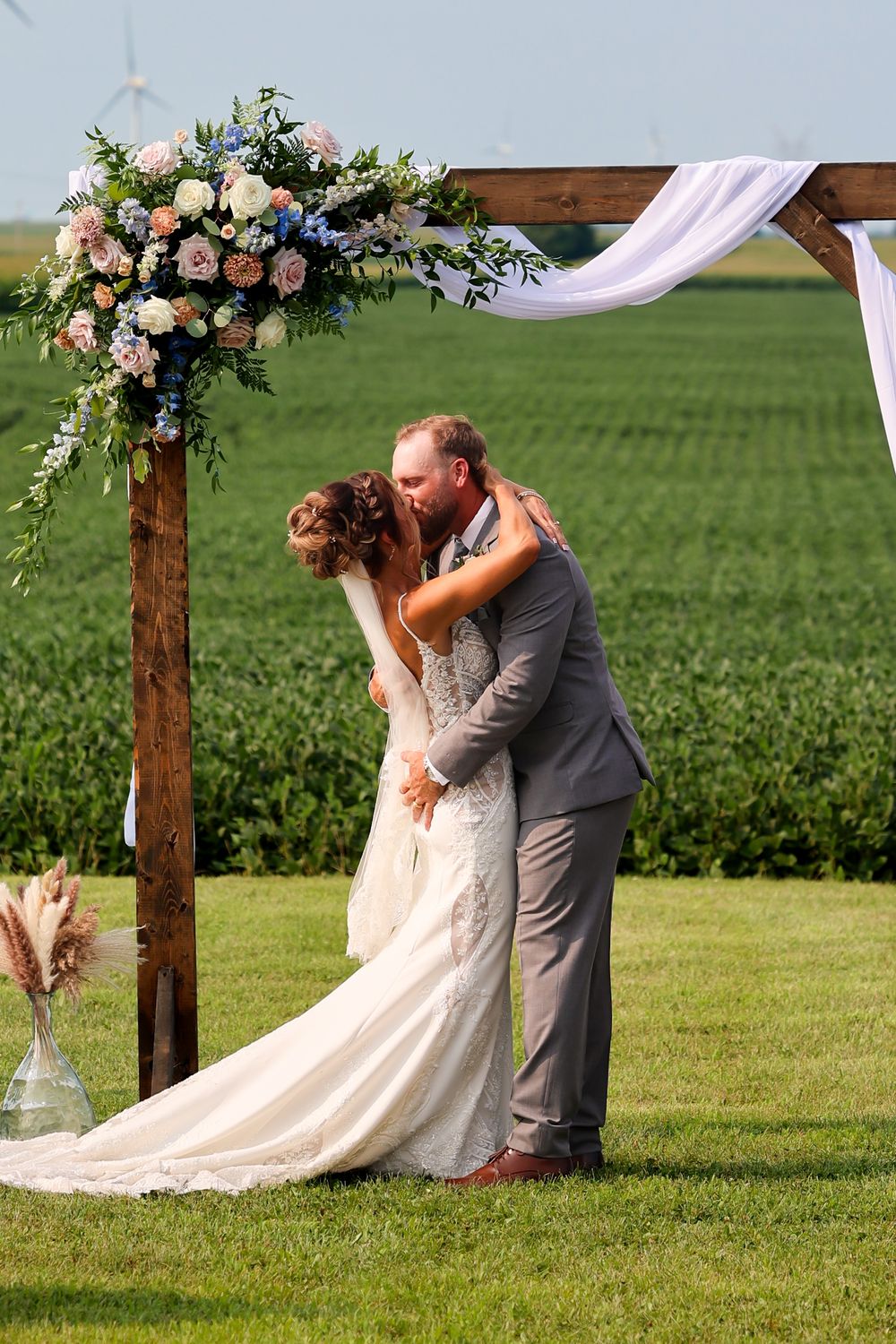 Bride and groom share first kiss under floral wooden arch with green cornfield backdrop.