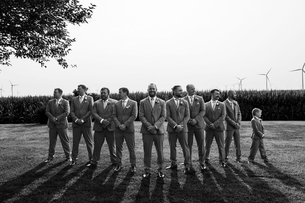 Groom and groomsmen in matching gray suits pose in black and white photo with wind turbines.