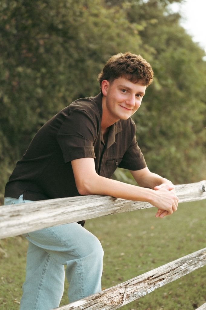 Young man in brown shirt and jeans leaning on a rustic wooden fence outdoors, smiling at the camera.