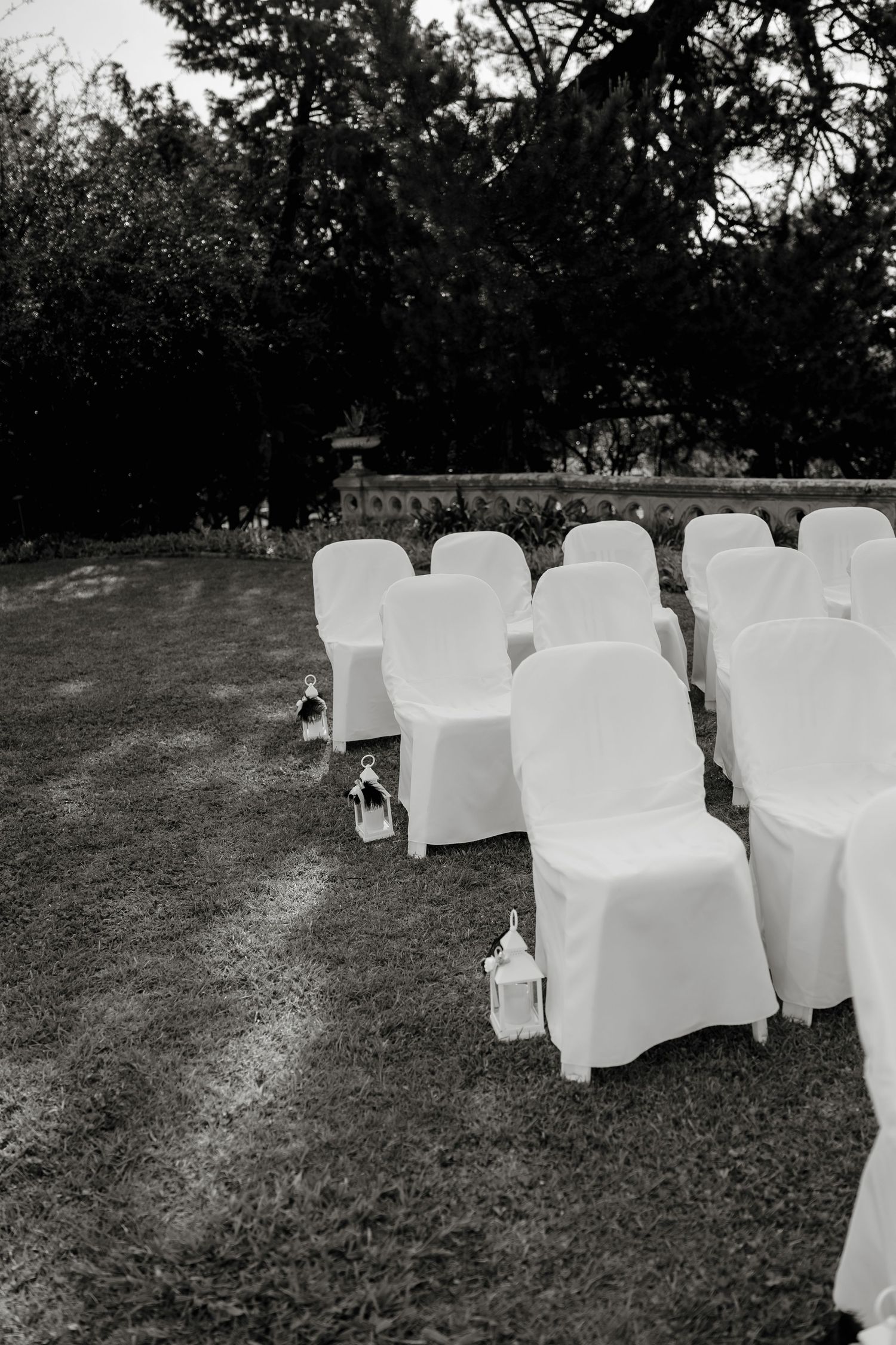 Black and white photo of white chair covers arranged in rows on grass for an outdoor wedding ceremony with lanterns.