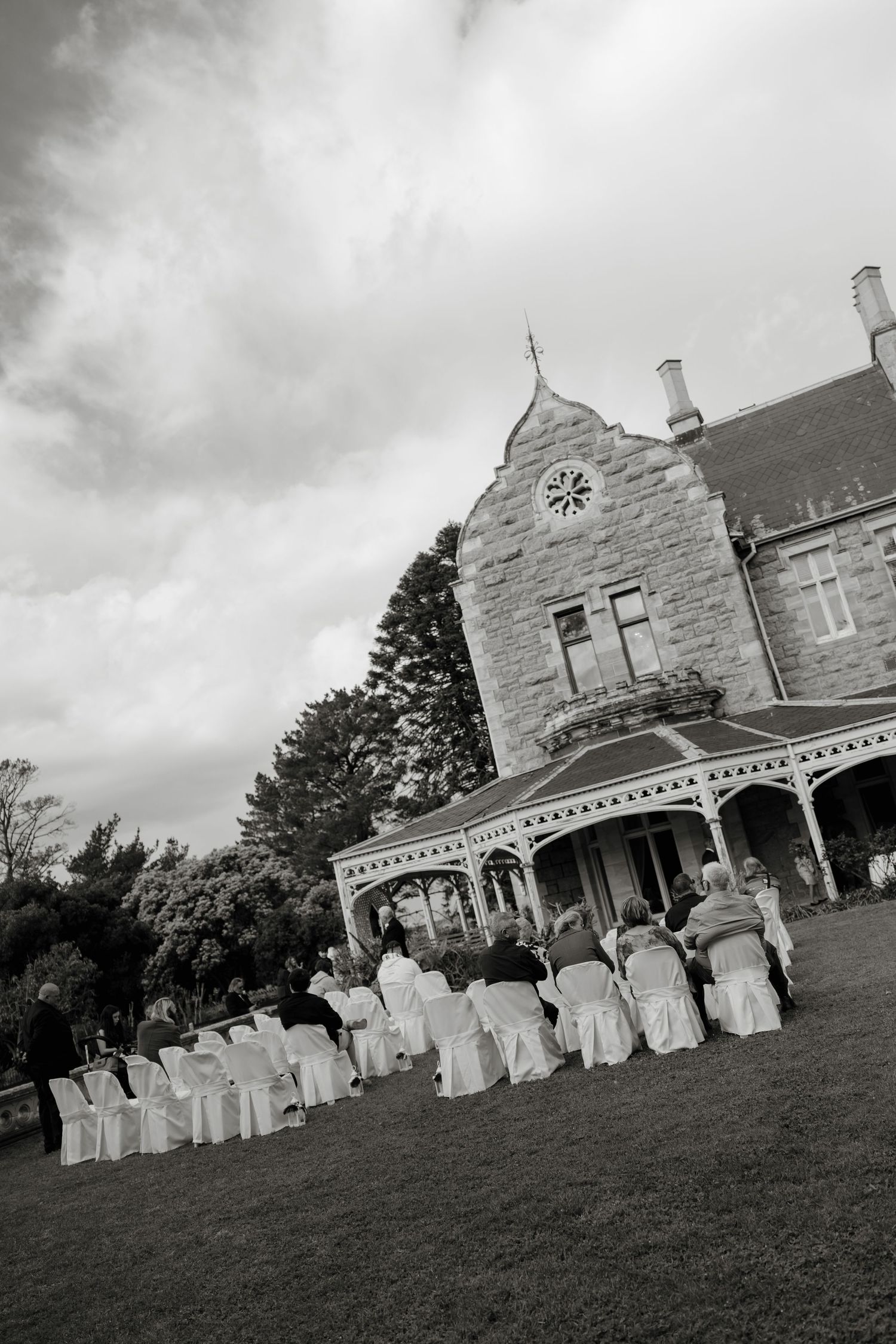 Black and white outdoor wedding ceremony on mansion lawn with white chair covers and historic stone building backdrop.