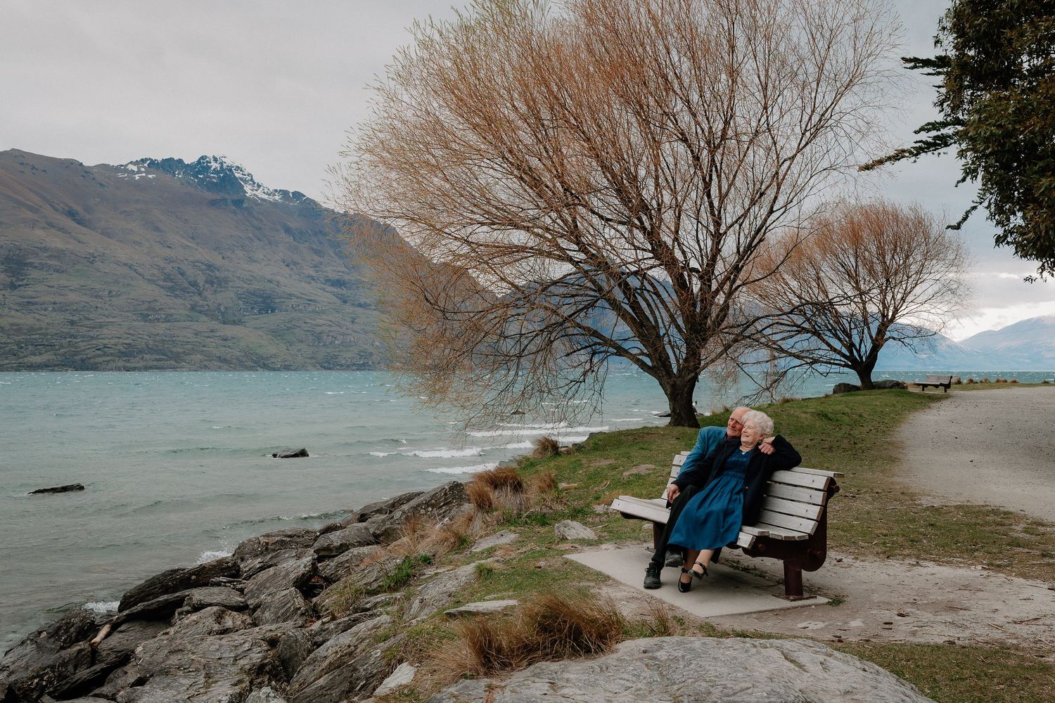 Elderly couple sitting on a wooden bench beside a rocky lake shore, with bare autumn trees and misty mountains in the background.