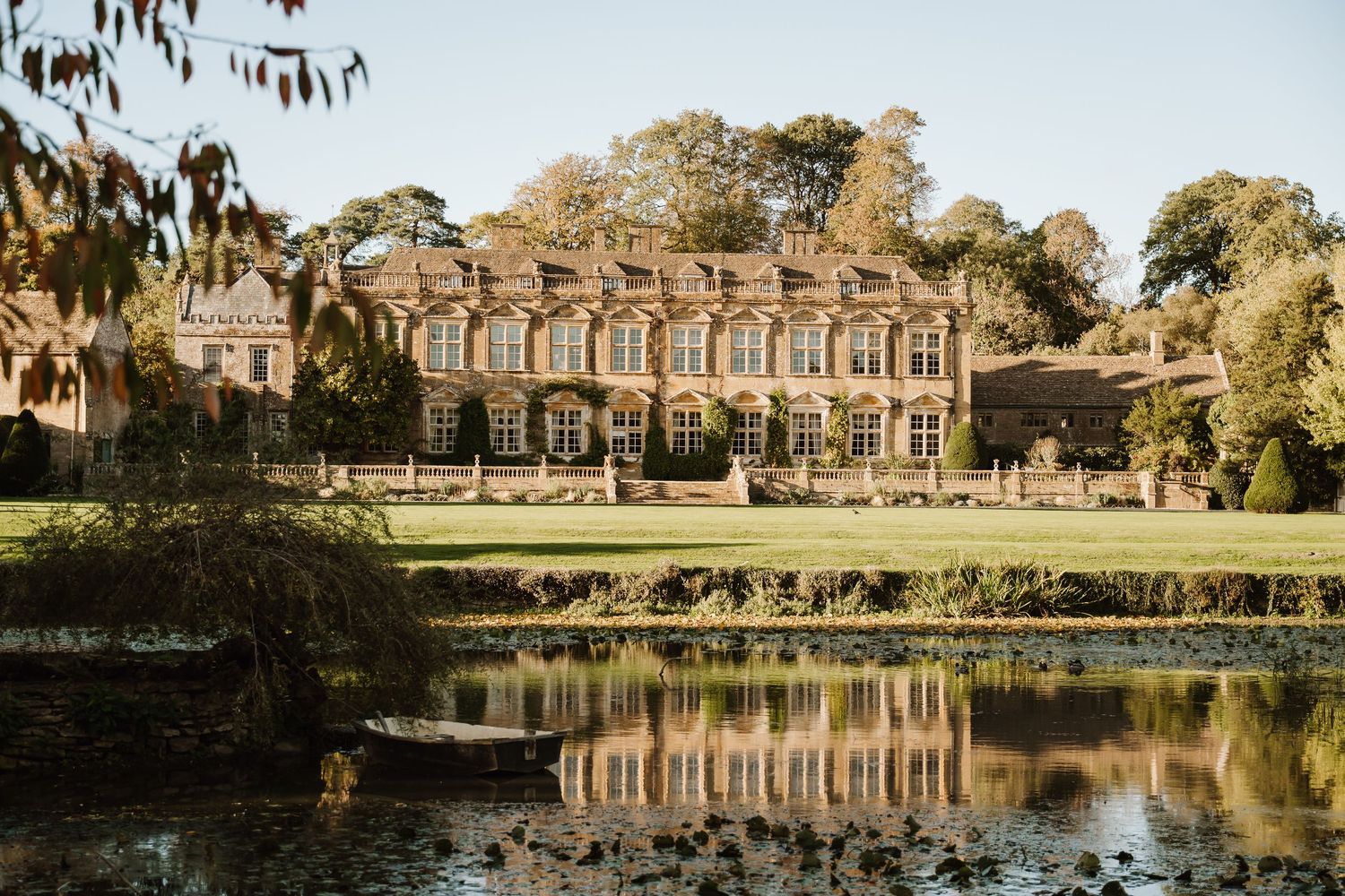 Historic stone manor house with tall windows and manicured lawns reflected in a calm pond, surrounded by mature trees on a sunny day.