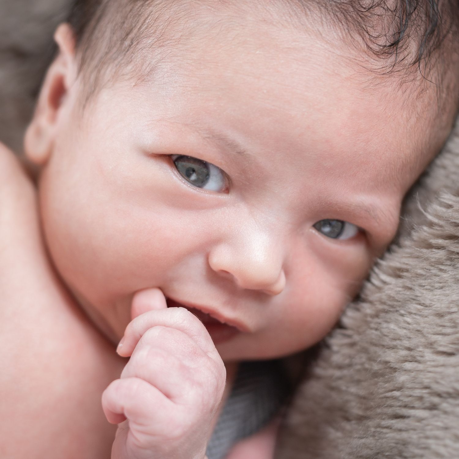 Close-up newborn portrait with blue-grey eyes, tiny fingers near the mouth and soft neutral textures.