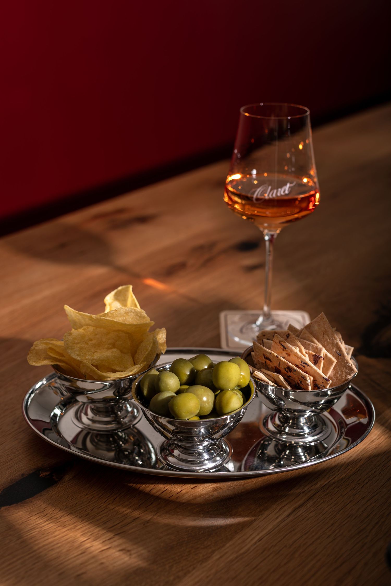 Silver serving tray with potato chips, green olives and crackers beside a glass of rosé wine on a rustic wooden table.
