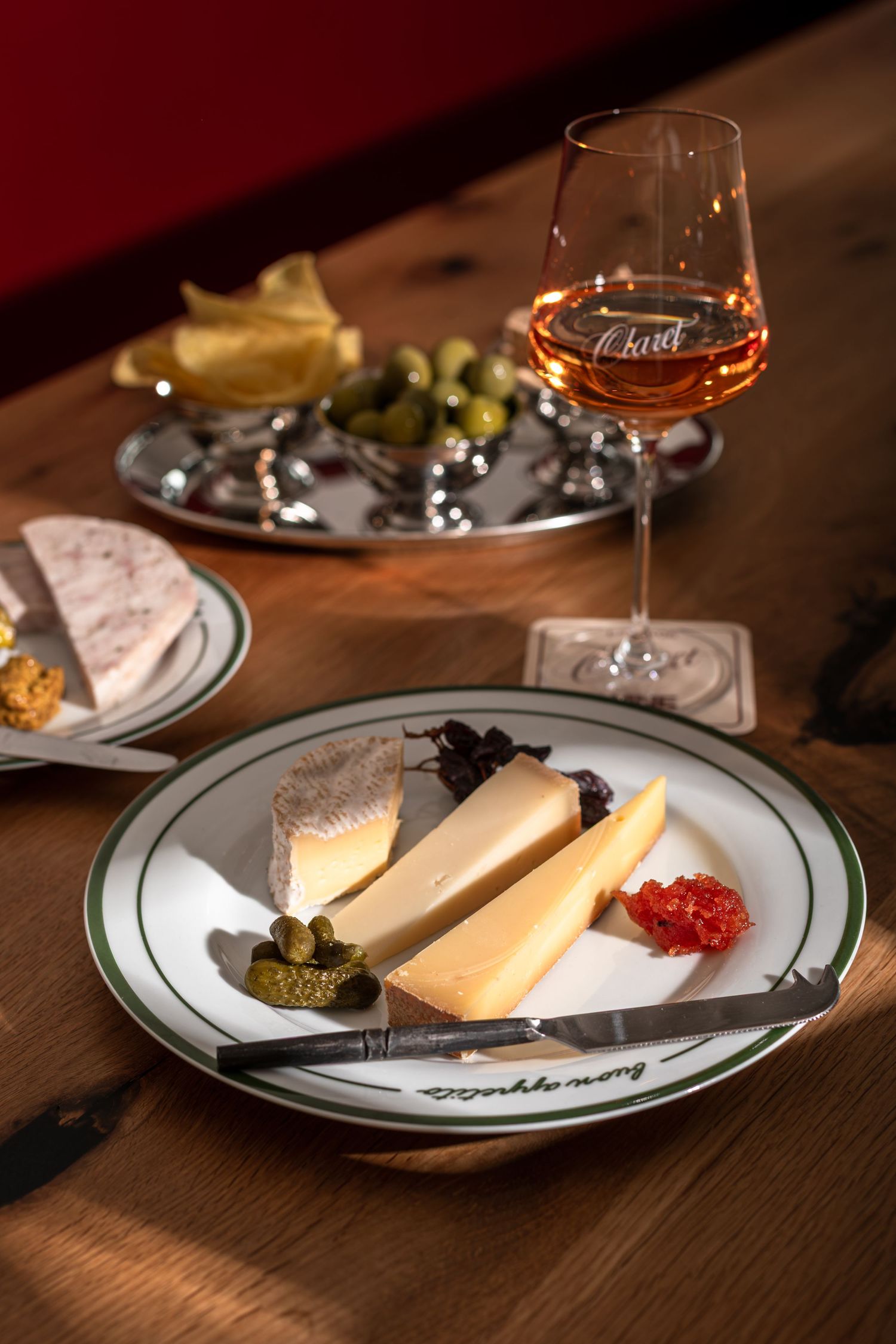 Cheese plate with sliced cheeses, cornichons and red relish beside a glass of rosé wine and a tray of olives on a wooden table.