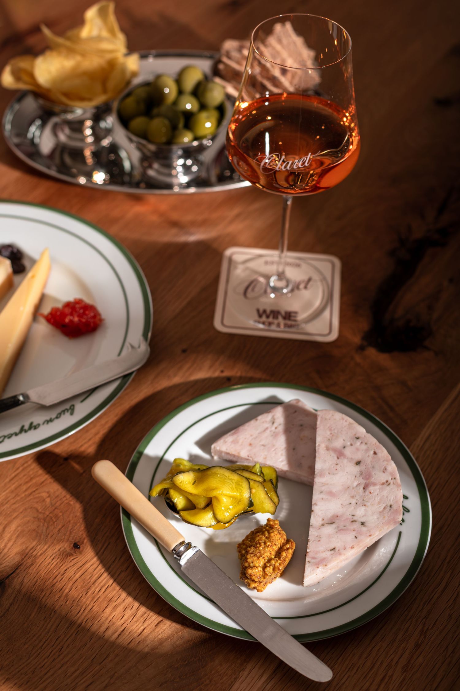 Glass of rosé wine on a coaster beside a plate of pâté with pickles and mustard, olives, chips and cheese on a wooden table.