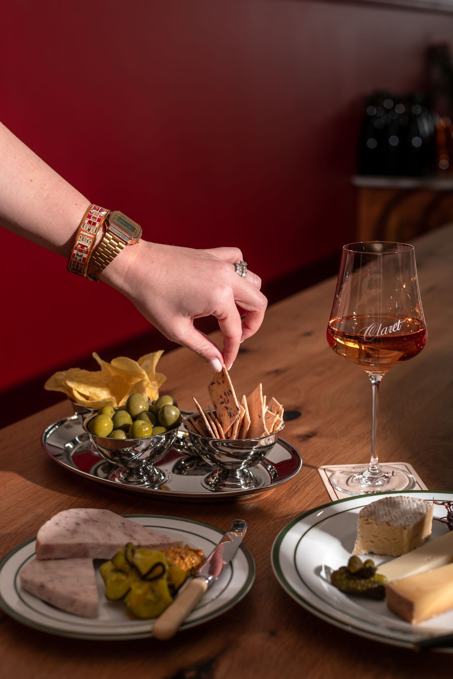 Hand reaching for a cracker from a silver dish beside green olives, a glass of rosé wine, and plates of cheese and pâté.