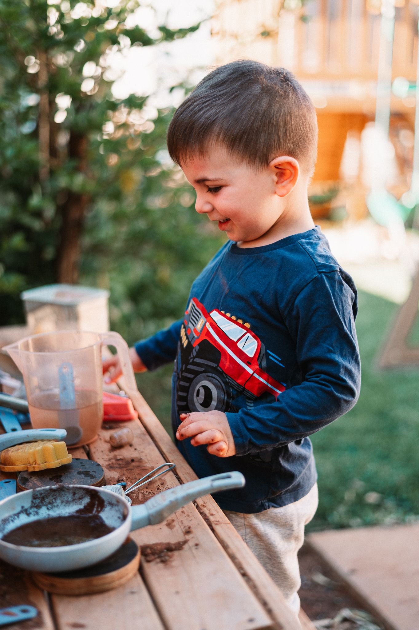 Young child in a navy sweater plays at a wooden outdoor mud kitchen in a sunlit backyard with fallen oranges scattered on the dirt.