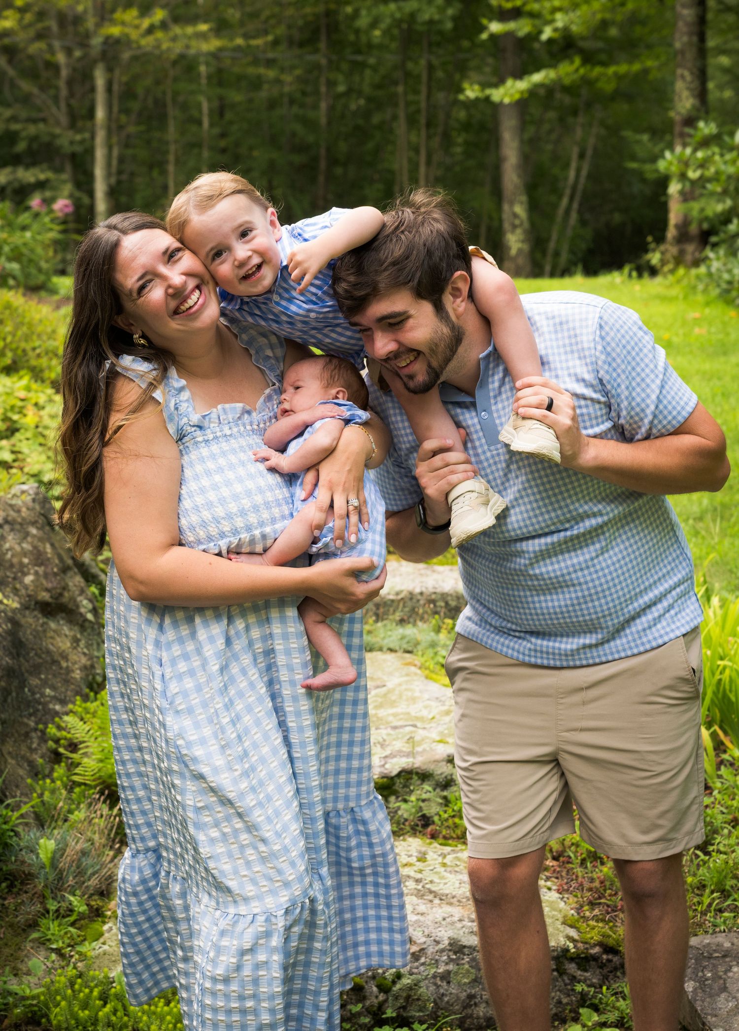 Smiling family in matching light blue gingham outfits holding two young children on a stone path surrounded by lush green woods.