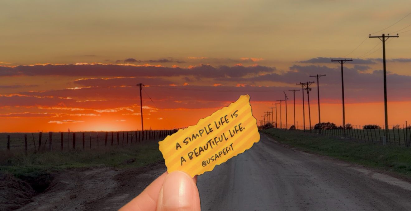 Hand holding a note saying 'a simple life is a beautiful life' against a stunning rural sunset road.