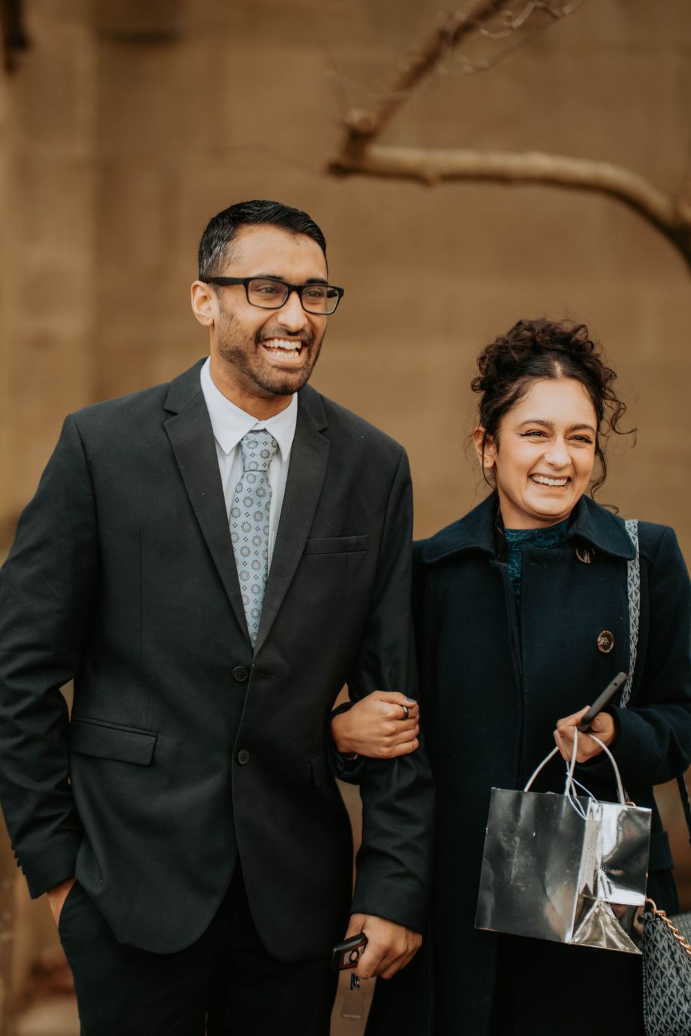 Smiling couple in formal attire walking together, man in suit with glasses, woman in navy coat holding gift bags.