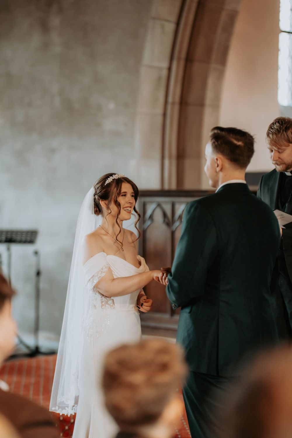 Bride smiling joyfully at groom during wedding ceremony in a church, wearing an off-shoulder white gown and veil.