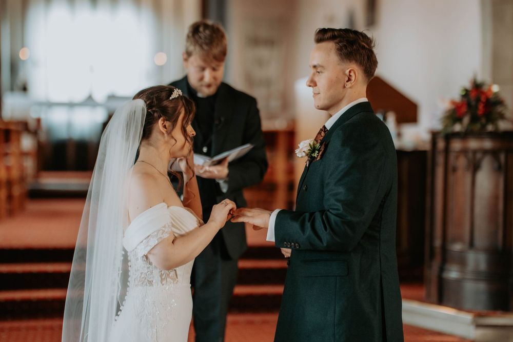 Bride placing ring on groom's finger during church wedding ceremony with officiant in background.