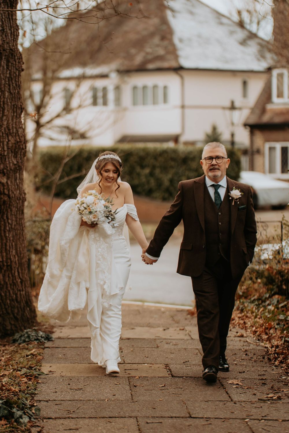 Bride in white lace gown walking hand in hand with father on suburban pathway.