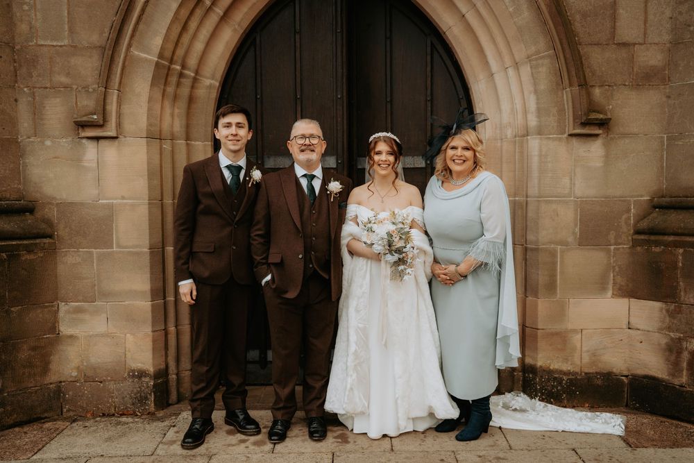 Wedding family portrait of four people posing before a stone church arched doorway.