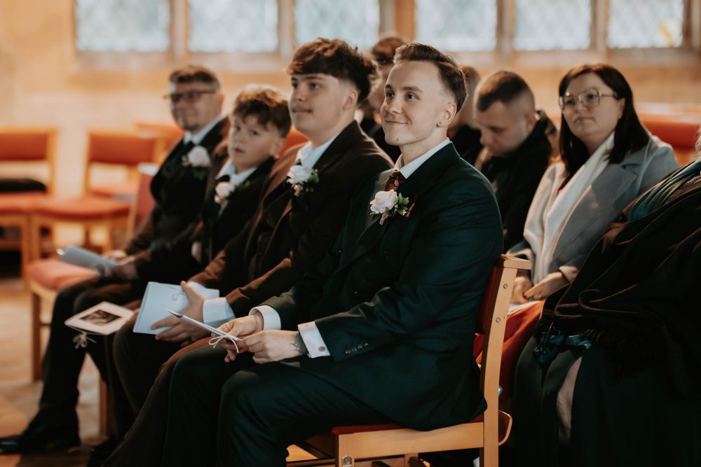 Groomsmen in dark suits with boutonnieres seated in wooden chairs at a wedding ceremony venue.