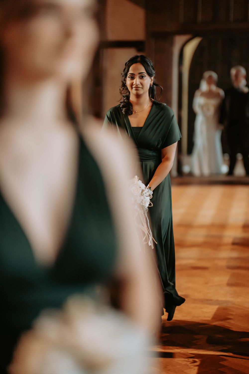 Bridesmaid in green gown holds white bouquet walking down aisle at elegant indoor wedding ceremony.