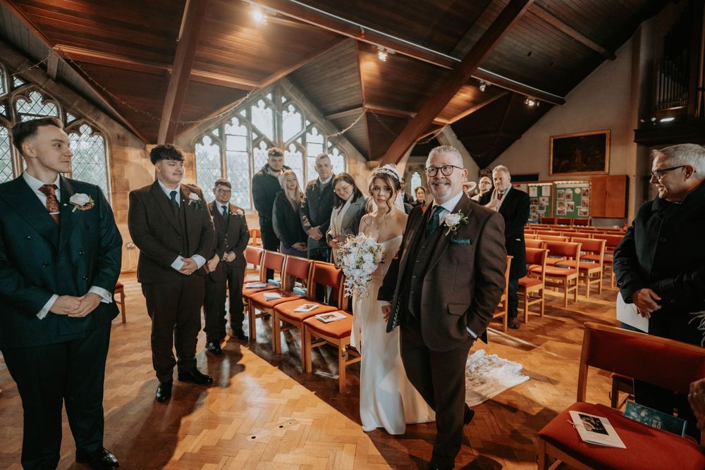 Bride walks down church aisle with father during wedding ceremony, guests standing in wooden chapel.