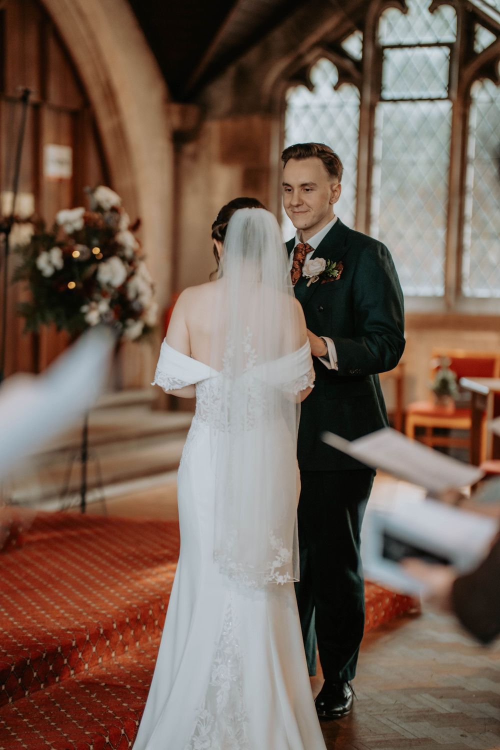 Groom in dark suit smiles at bride in white off-shoulder gown during church wedding ceremony.