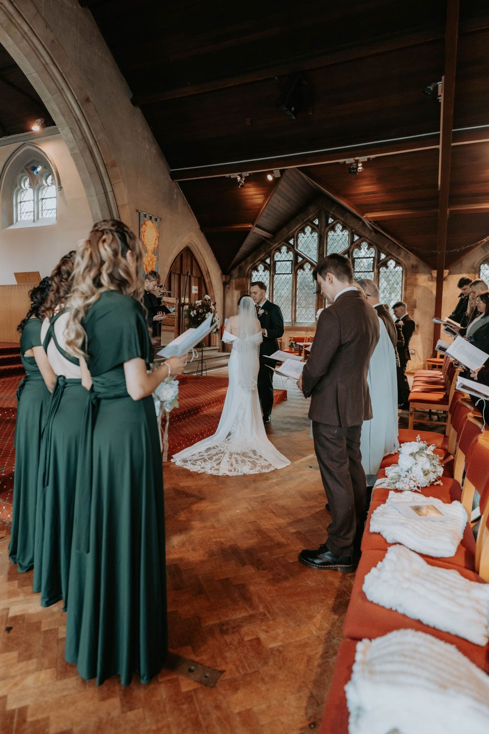 Bridesmaids in emerald green dresses watching couple exchange vows at church altar.