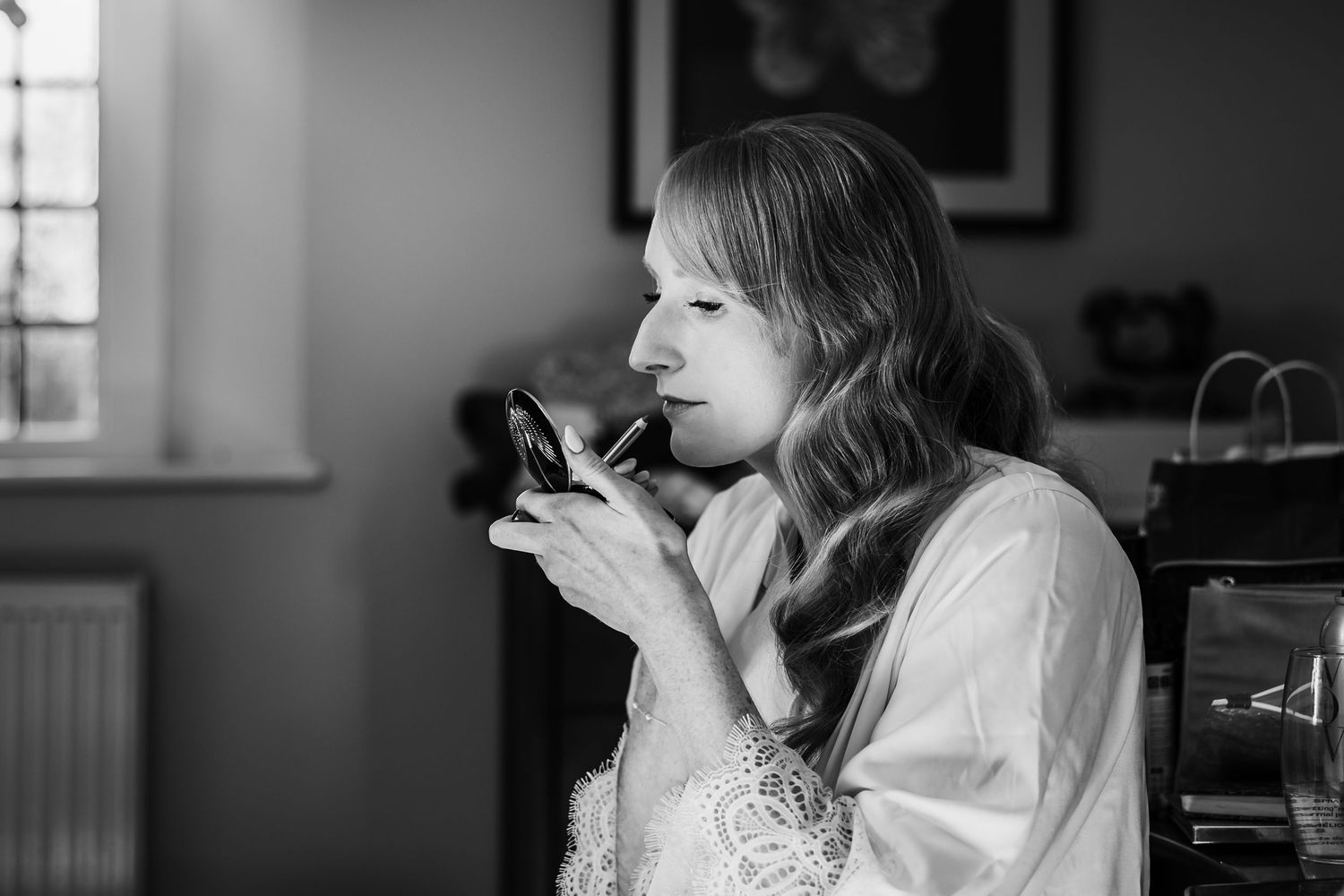 Black and white photo of a woman in a lace-trimmed robe applying makeup while looking into a small handheld mirror by a window.