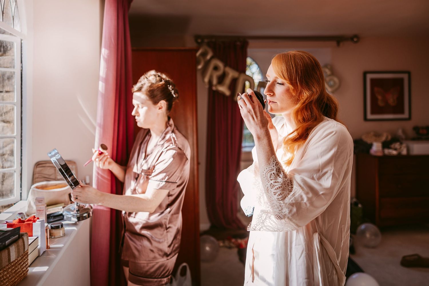 Bride in a white robe and a bridesmaid in a satin robe get ready by a sunlit window on the morning of the wedding.
