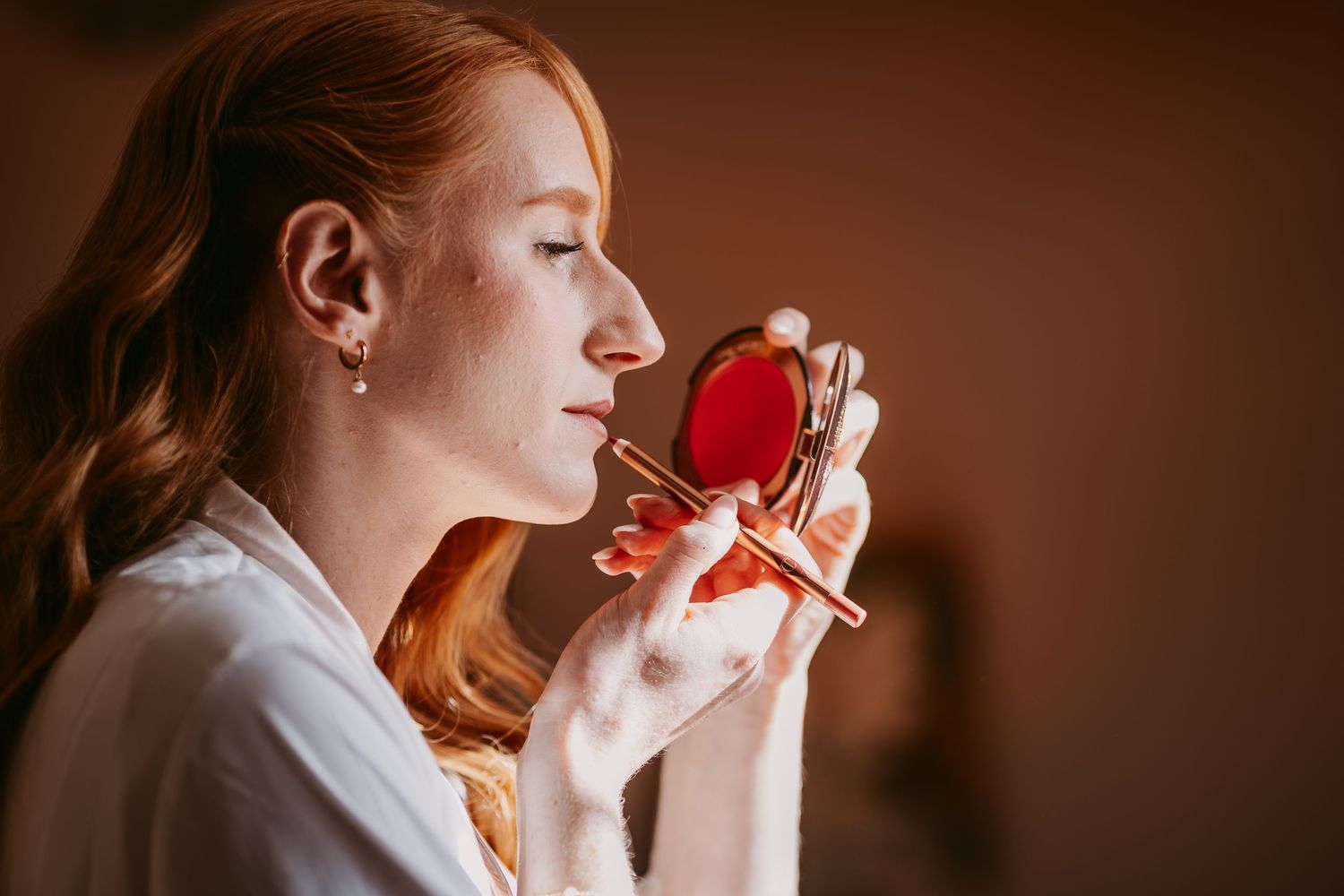Red-haired bride in a white robe applies lipstick while looking into a compact mirror during bridal preparations.