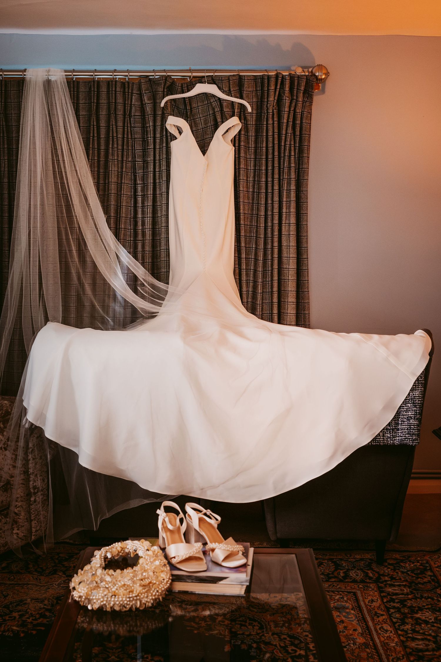 White wedding gown and long veil hang from a curtain rail above bridal sandals and a floral hair wreath on a table.