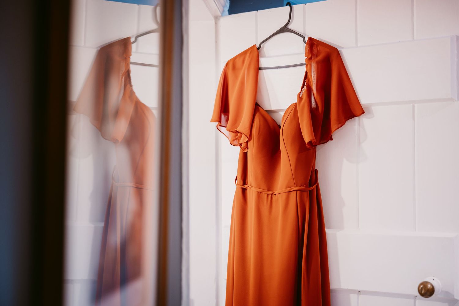 A rust-orange bridesmaid dress with flutter sleeves and a tie waist hangs on a white door, reflected in a nearby mirror.