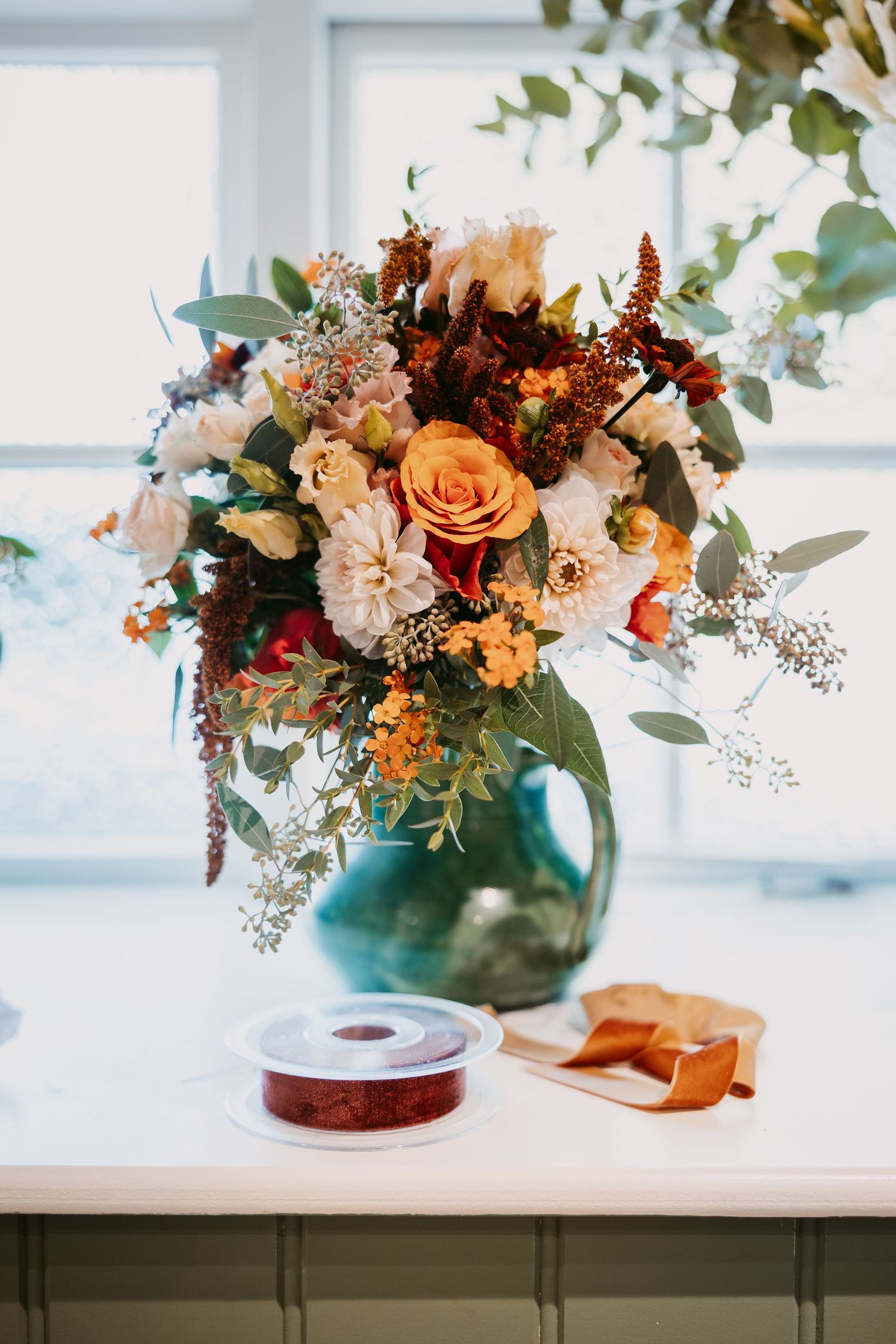 Autumn wedding bouquet of roses, dahlias and foliage in a green glass vase on a windowsill beside a copper ribbon spool.