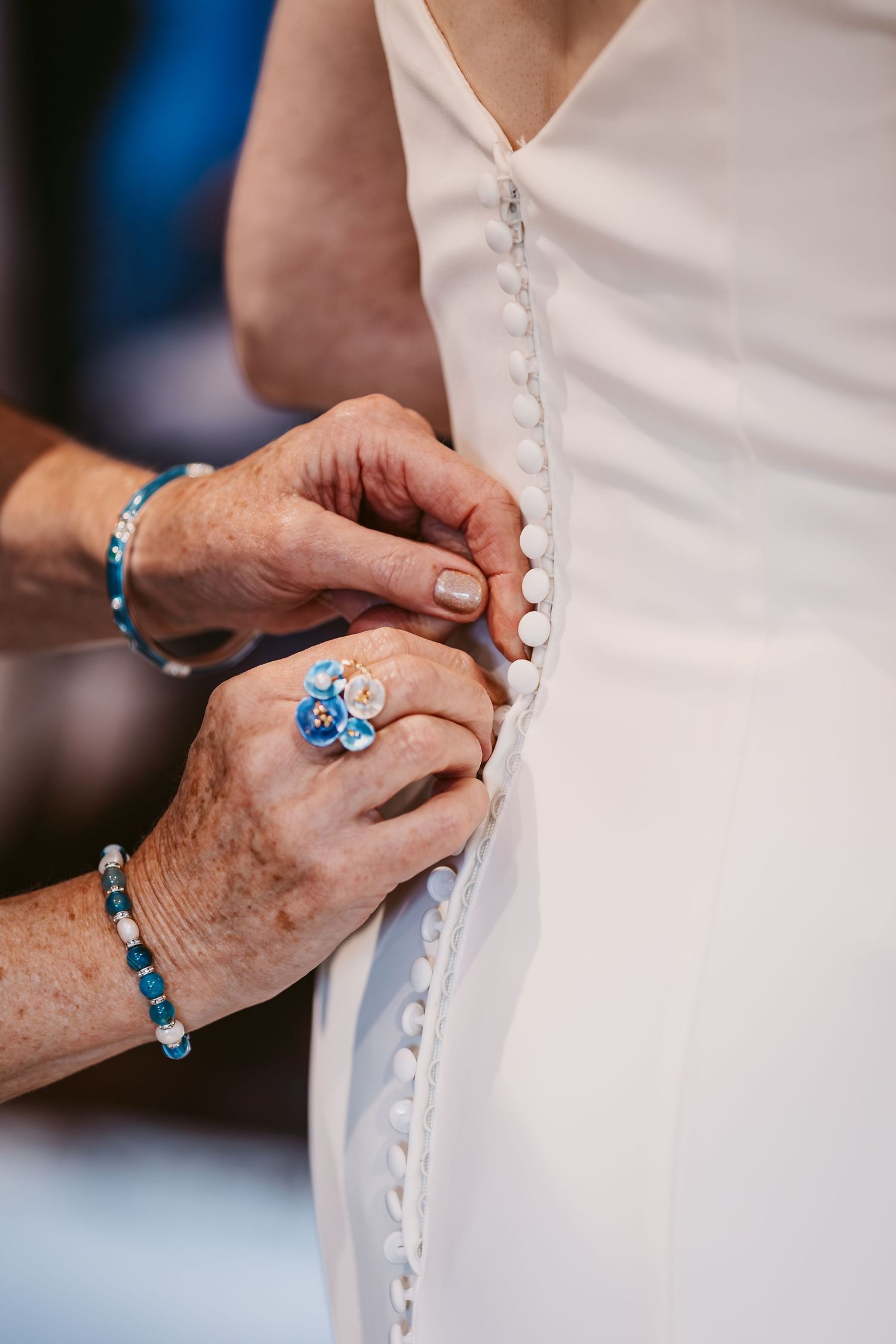 Elderly hands with a blue floral ring and beaded bracelets carefully fasten the small fabric-covered buttons on the back of a wedding dress.
