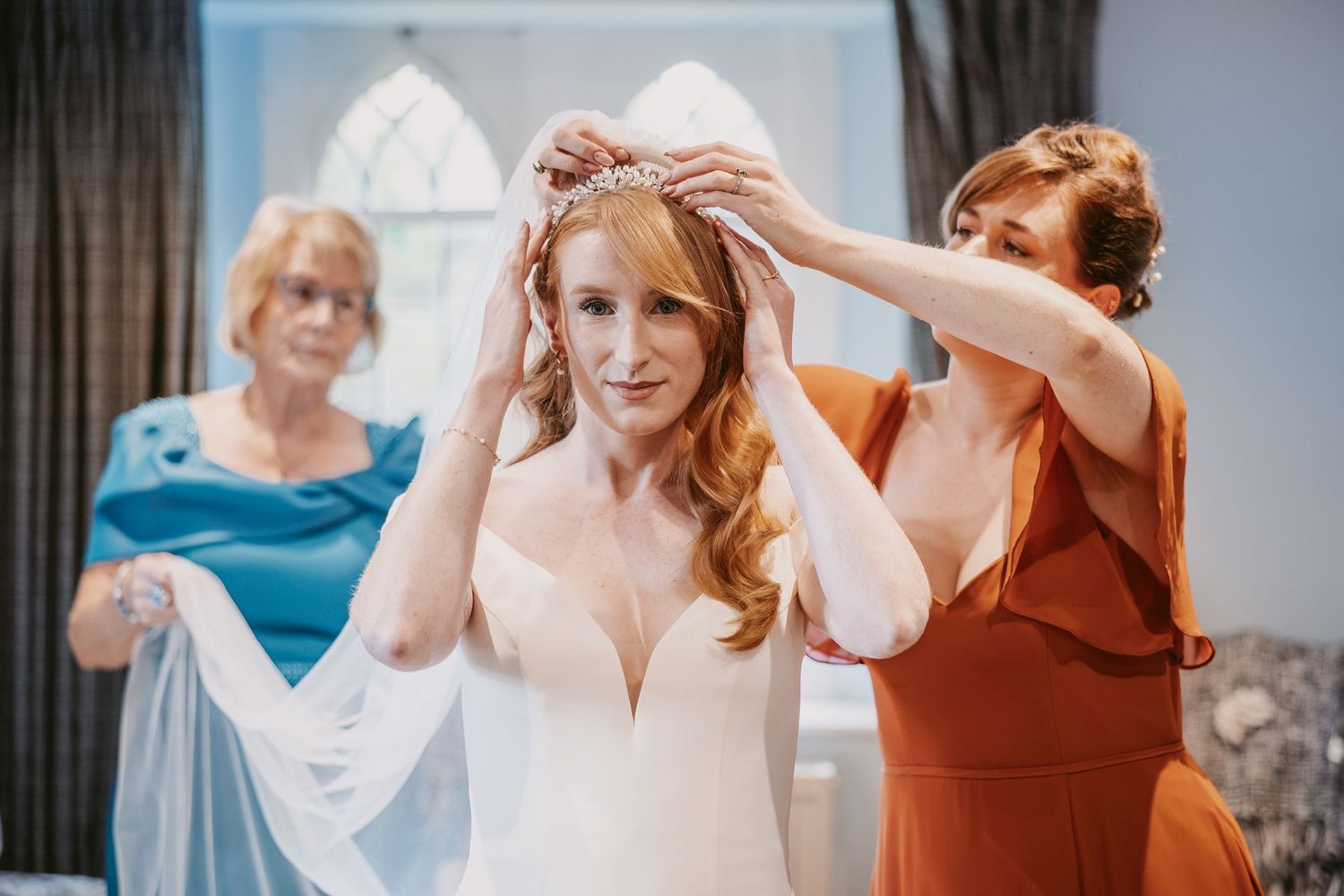 A bridesmaid in a rust dress helps a red-haired bride place a tiara while an older woman in teal holds the veil nearby.