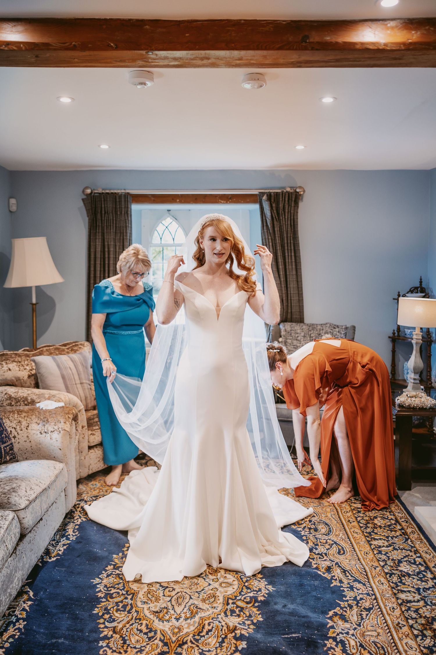 Bride in a fitted white wedding dress has her long veil adjusted by two women as she gets ready in a cozy room.