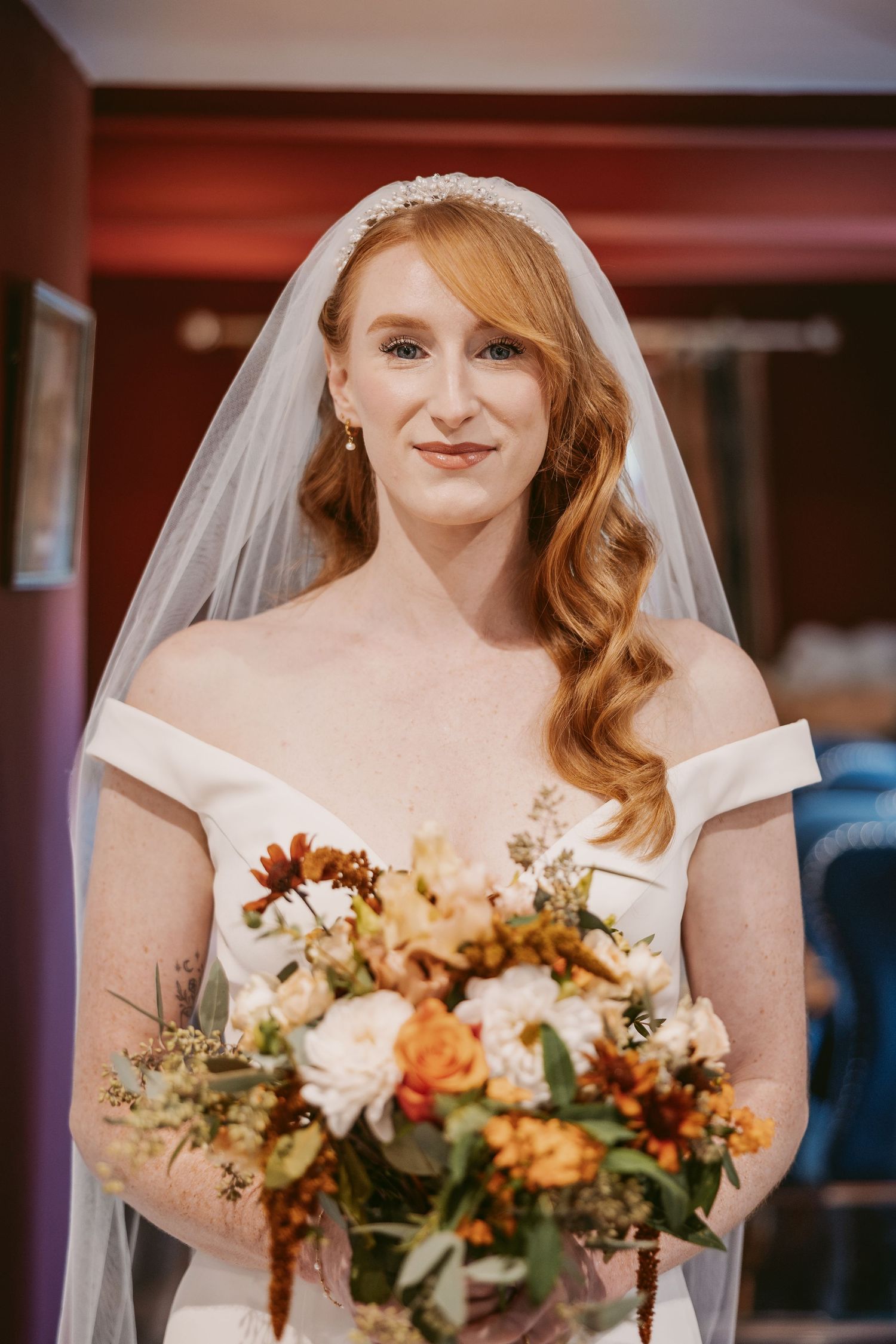 A smiling red-haired bride in an off-the-shoulder white gown, veil and tiara holds an autumn bouquet of orange and cream flowers.