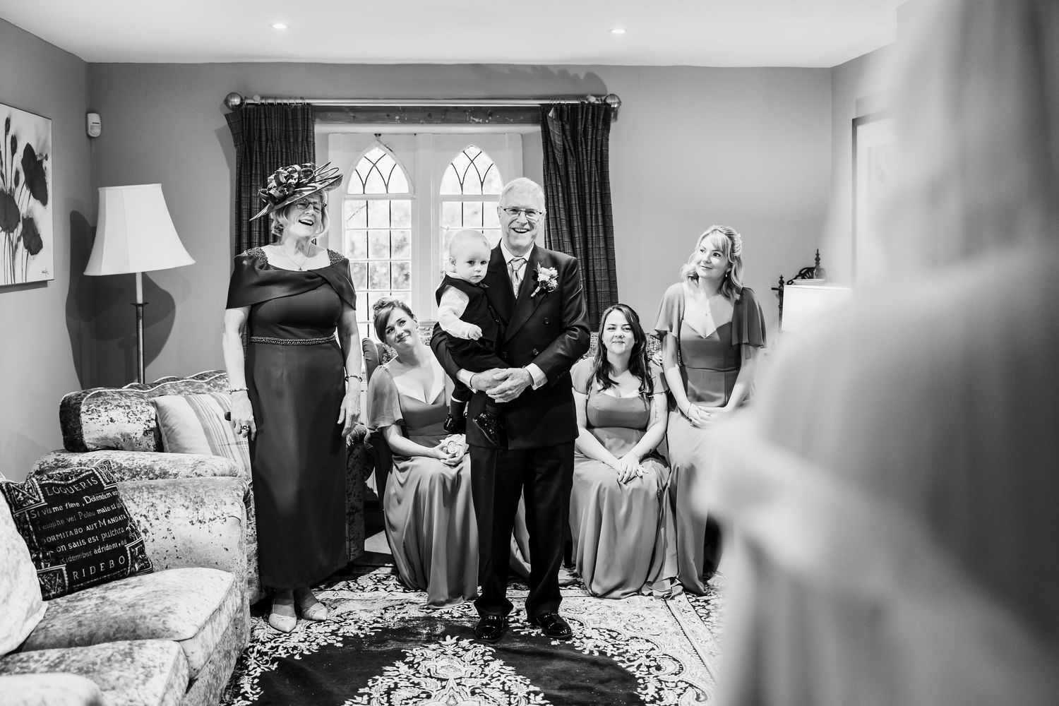 Black and white photo of a father holding a toddler as bridesmaids and a woman in a fascinator smile in a cozy room.