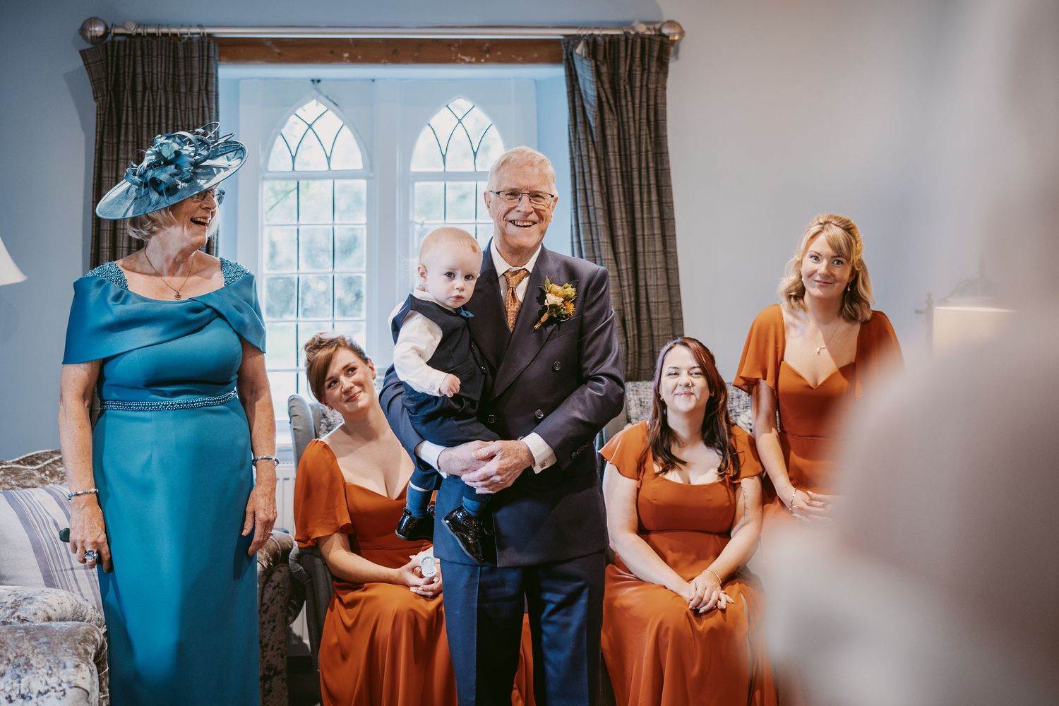 Father of the bride holds a toddler while smiling bridesmaids in rust dresses and a woman in a teal dress and hat look on.