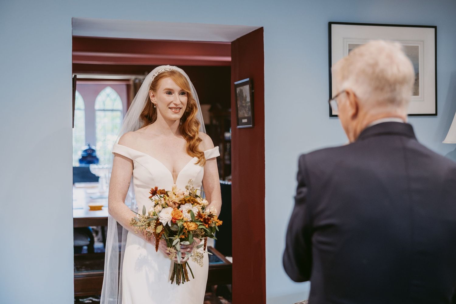 A bride in a white off-shoulder gown holds an autumn bouquet and smiles at an older man in a dark suit in a blue-walled room.