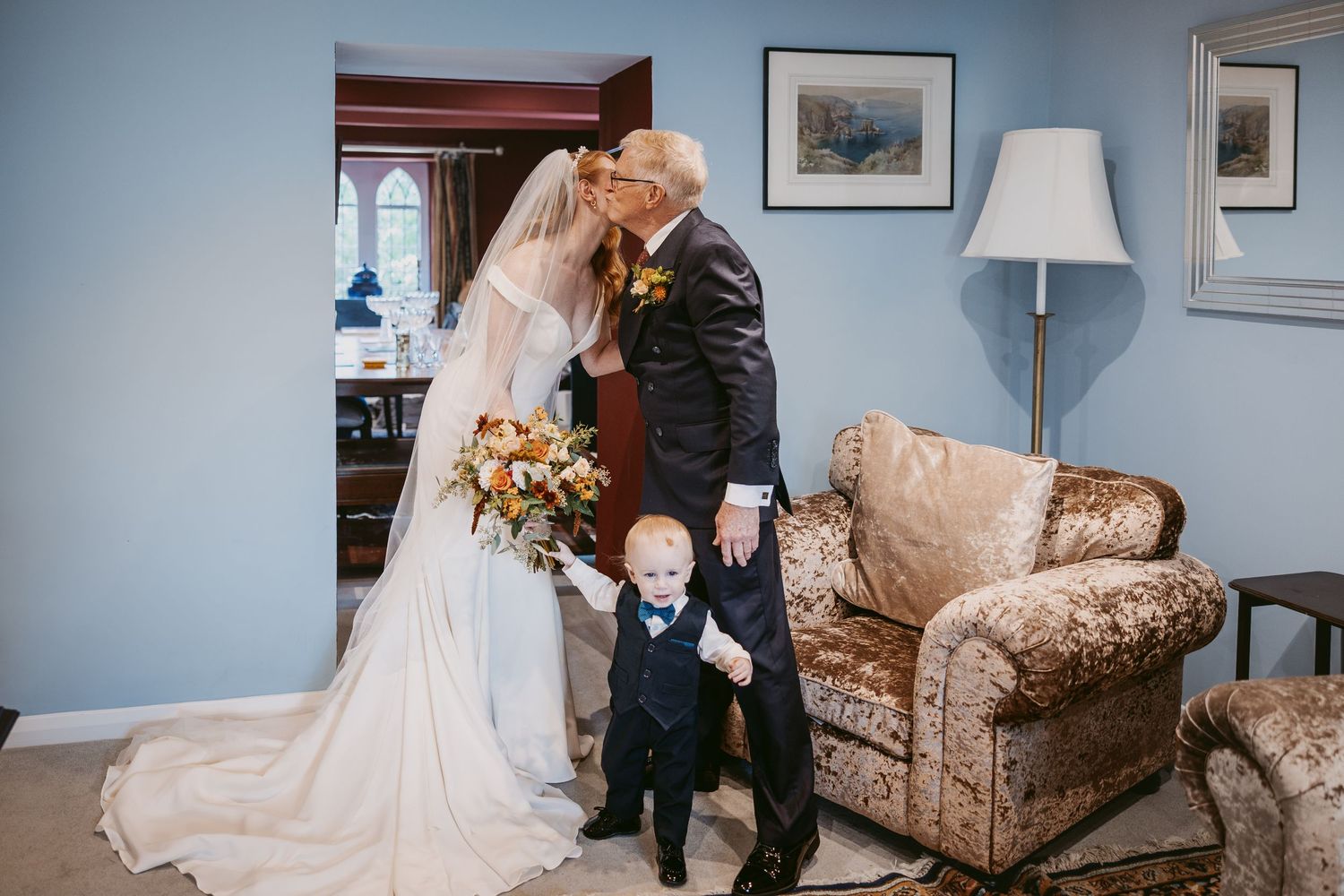 Father of the bride kisses the bride holding a bouquet as a young page boy in a bow tie reaches up beside them.